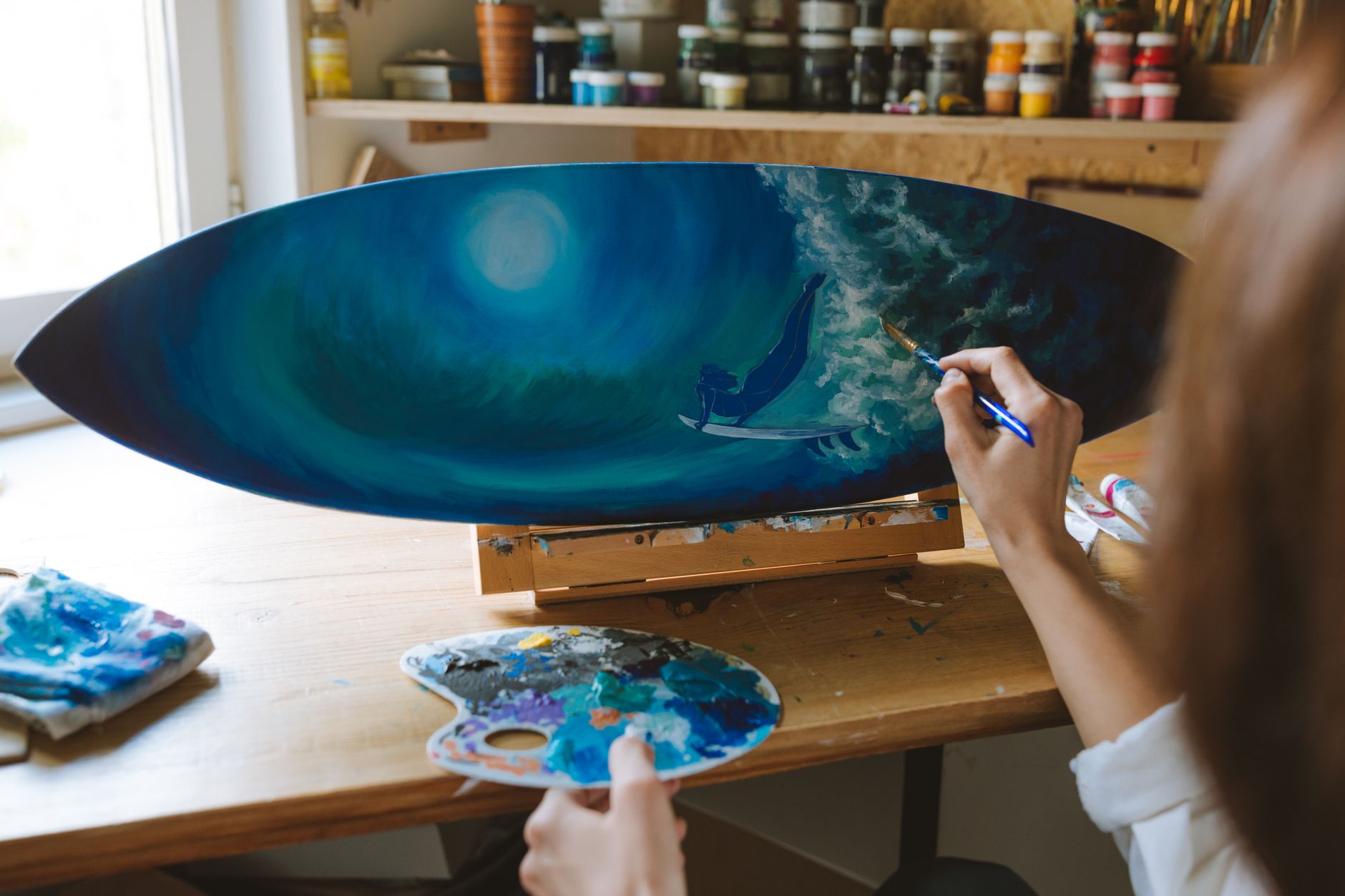 Artist painting a surf scene on a surfboard, showcasing artistic creativity in a sunlit studio in Carlsbad, CA